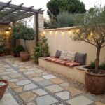 Narrow outdoor stone pathway with pebble accents flanked by raised planters filled with olive trees, shrubs, boxwoods, and terracotta pots, a cushioned bench against the wall, and wall-mounted lights along beige stucco exterior.