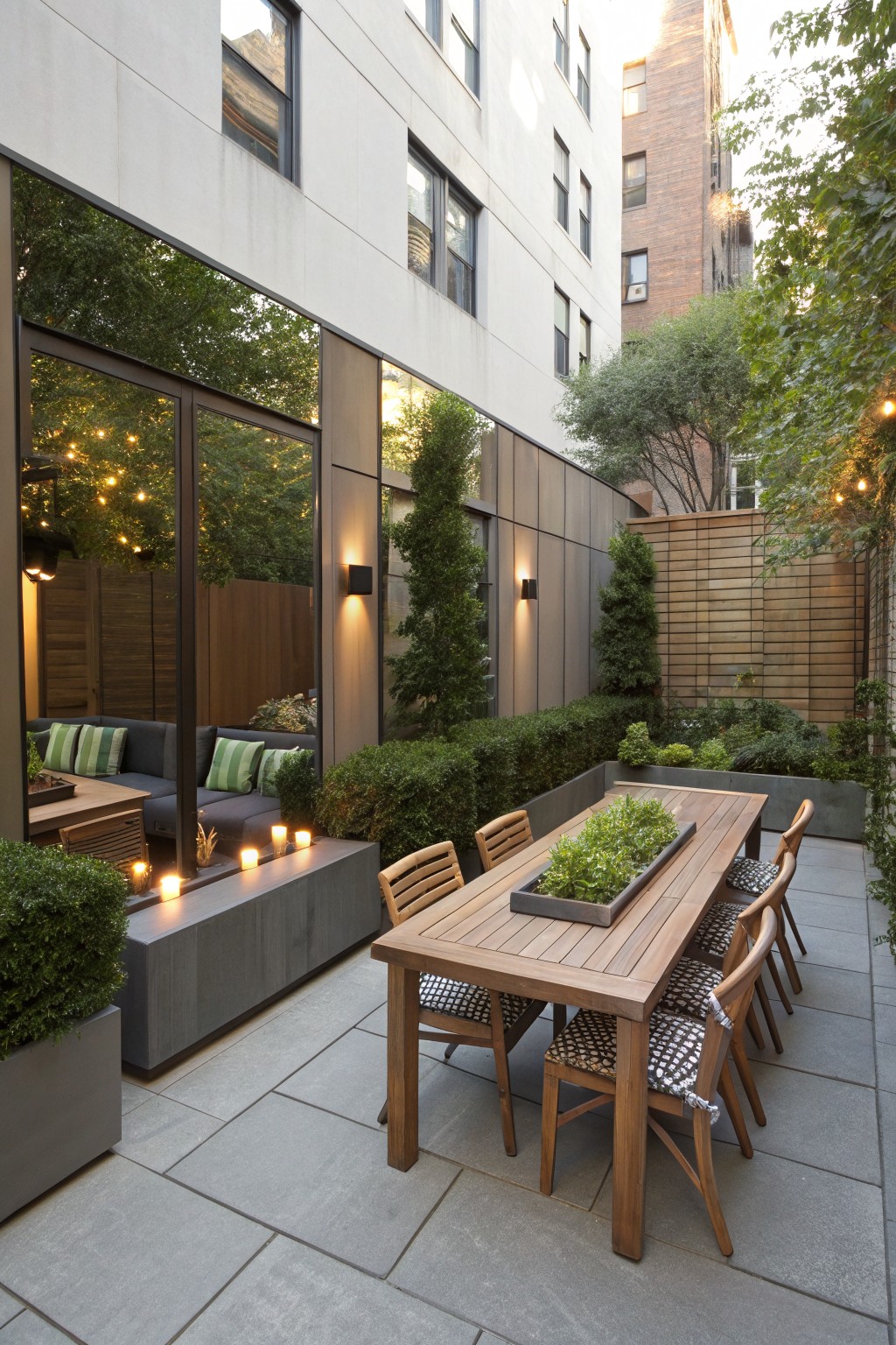 Small urban backyard patio featuring a wooden dining table with chairs, lounge seating area, potted plants and hedges, concrete fire bench with candles, string lights, and large reflective glass walls reflecting greenery against surrounding buildings.