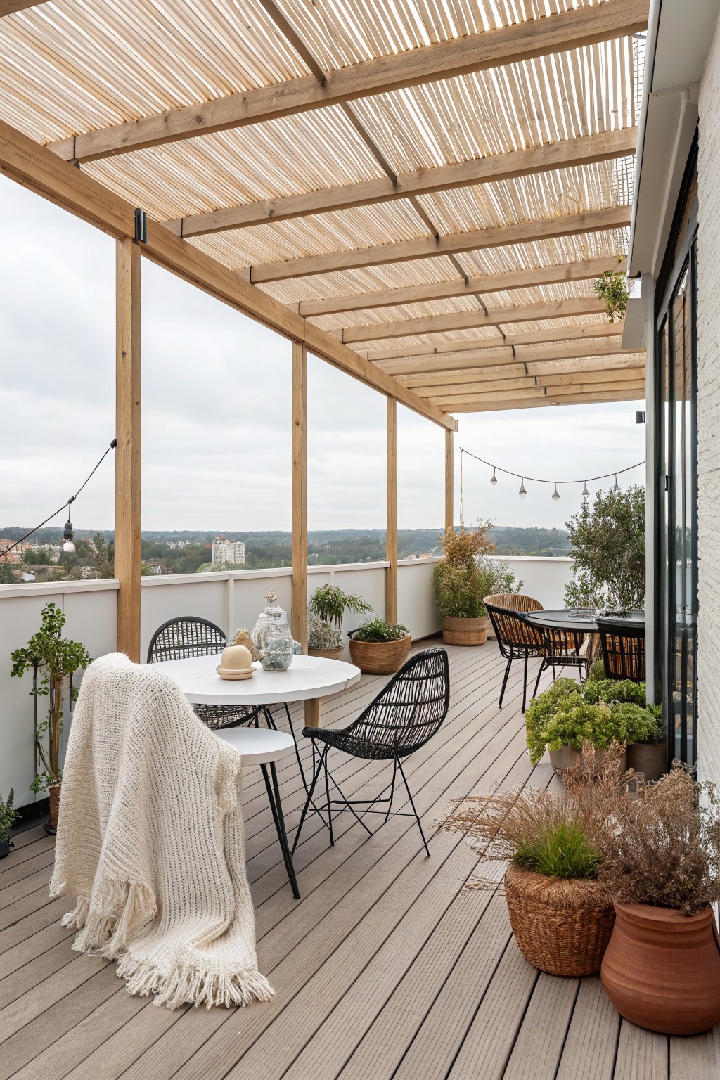 A modern rooftop deck with a wooden slatted pergola overhead, round table and chairs, potted plants, and a white blanket draped over one seat.