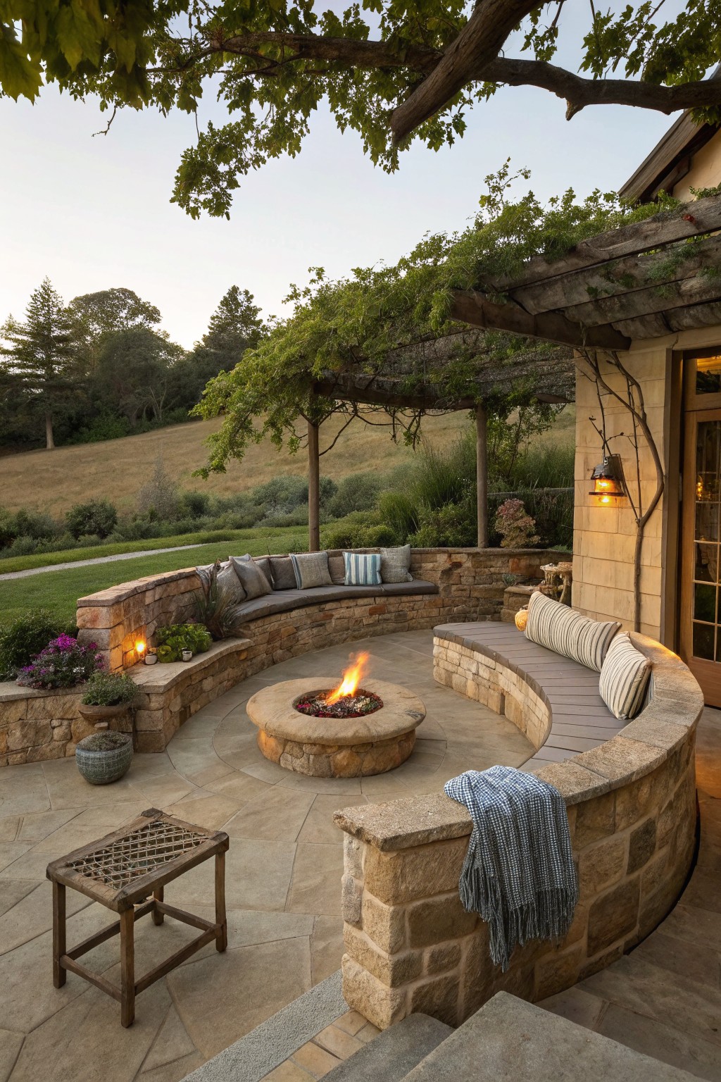 A circular stone fire pit on a patio is surrounded by curved built-in seating with cushions under a wooden pergola covered in vines.