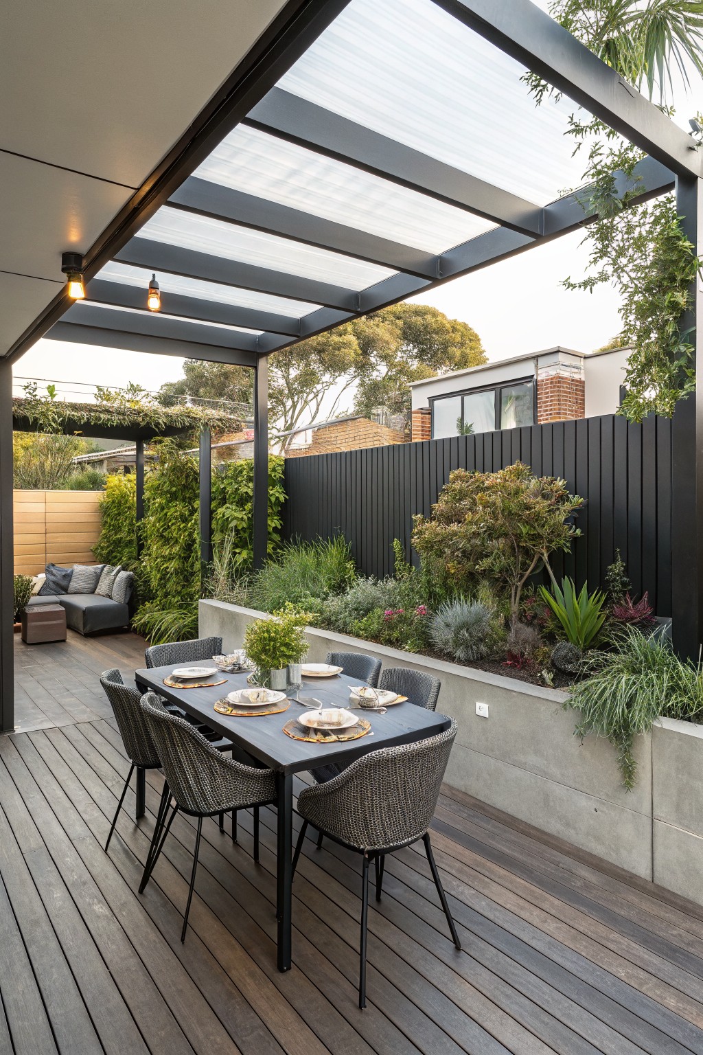 A modern dark-framed pergola with translucent and slatted panels covers a dining table and woven chairs on a wooden deck next to a concrete planter with shrubs and a black fence.