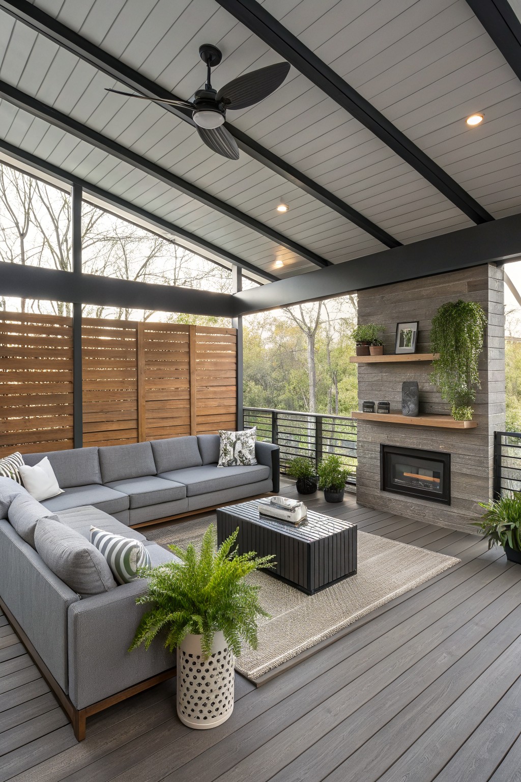 A covered outdoor living area with gray sectional sofas, a dark coffee table, a stone fireplace wall, a ceiling fan, and large windows overlooking trees.