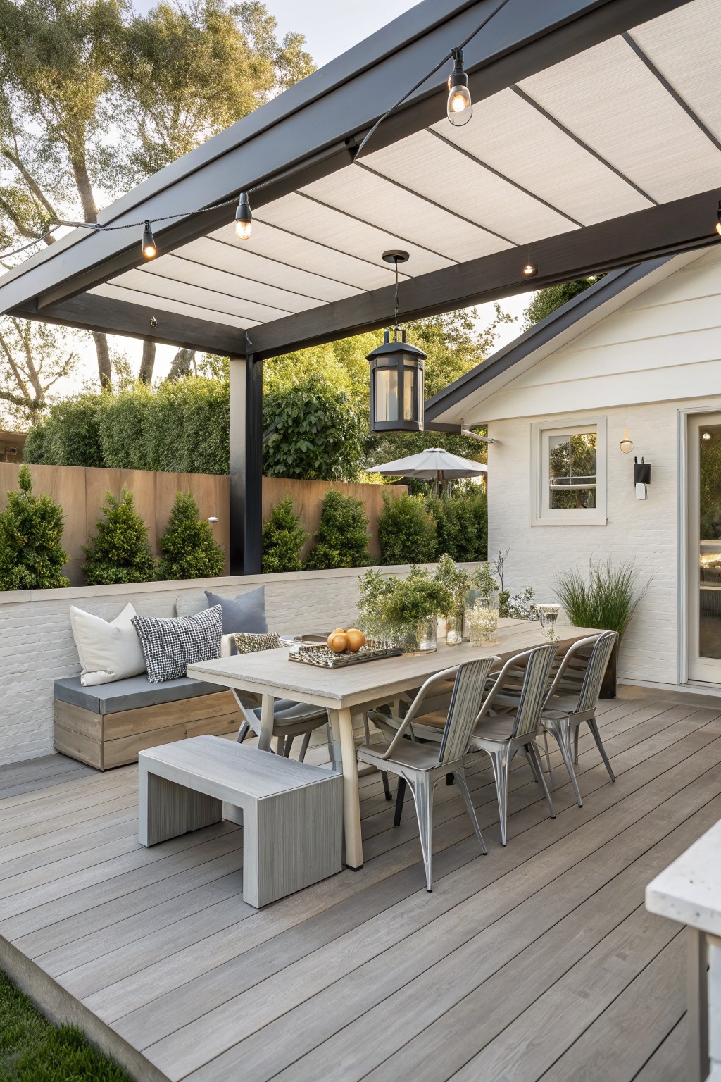 Outdoor deck dining area with a long table, metal chairs, built-in bench seating, and overhead cover with string lights.