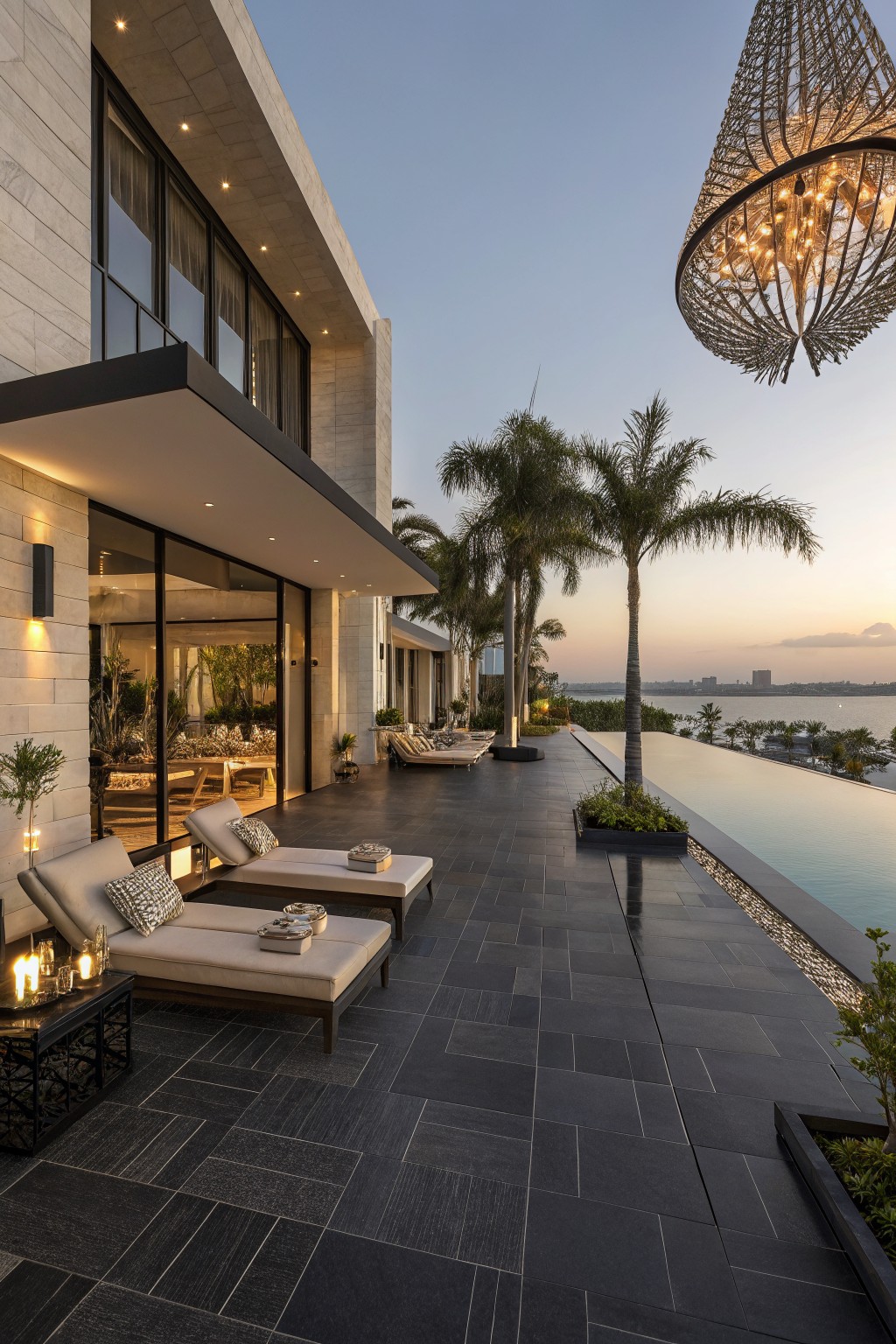 Modern outdoor patio with dark stone tiles, multiple lounge chairs, an infinity pool, and palm trees at dusk.