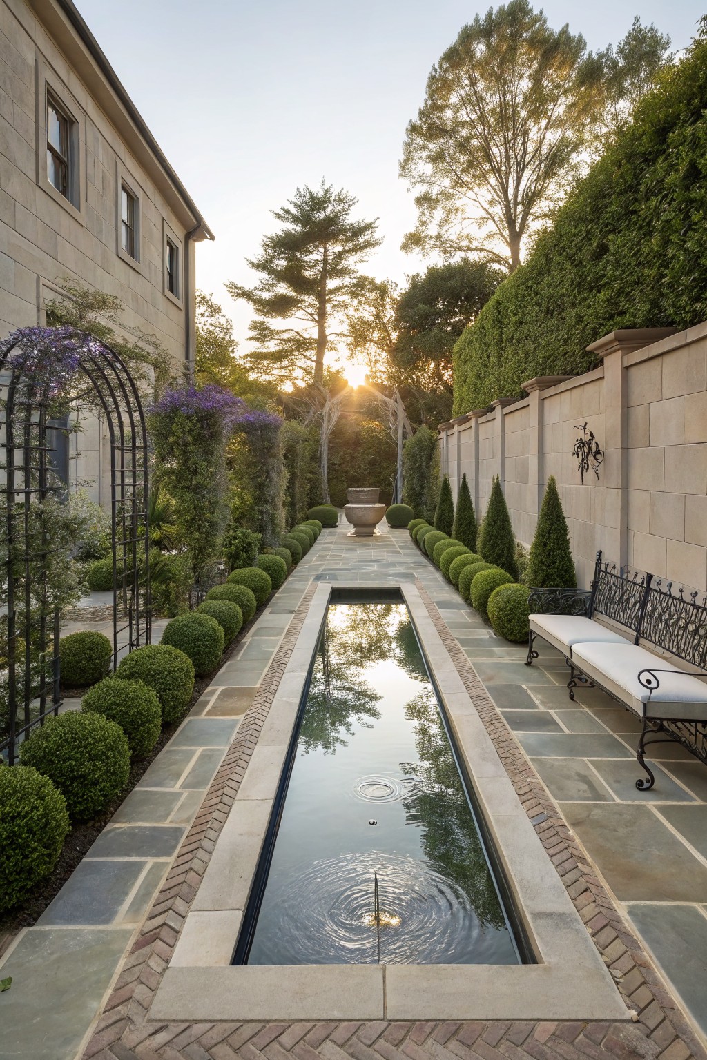 A long rectangular reflecting pool runs down the center of a narrow backyard lined with stone paths and rows of rounded shrubs.