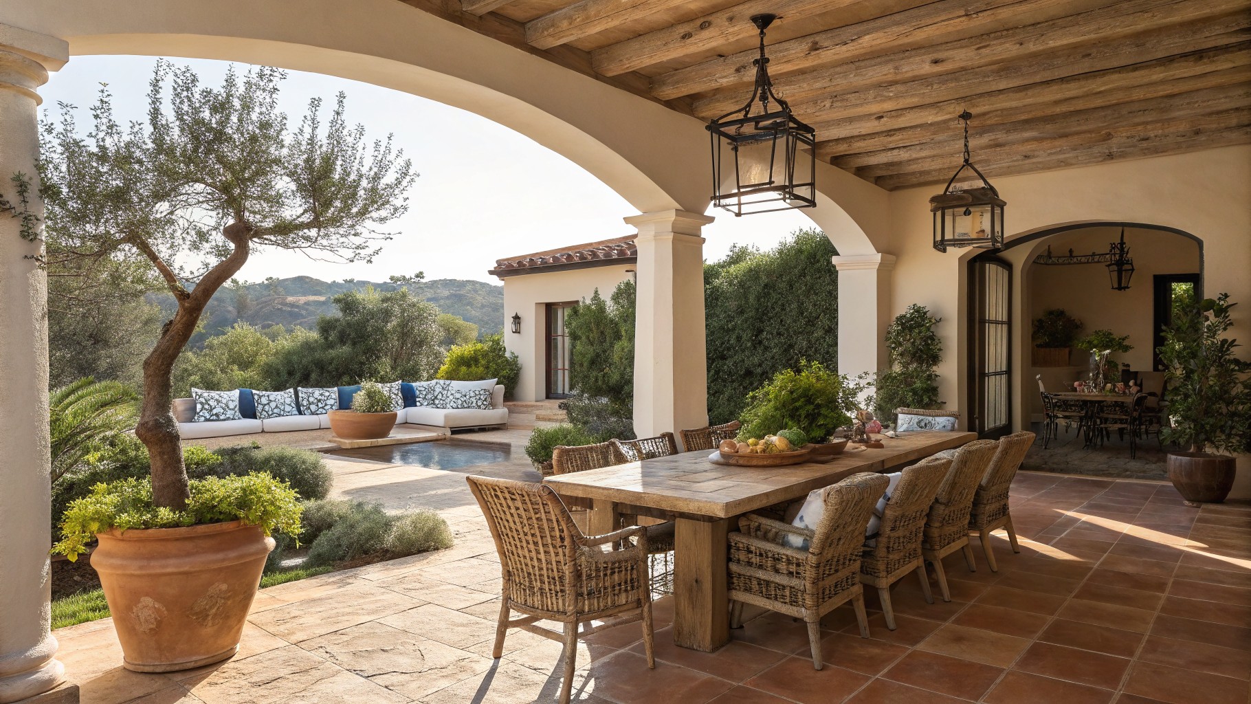 A covered patio featuring a large wooden dining table surrounded by woven chairs, a hanging metal lantern, and potted plants under exposed wood beams.