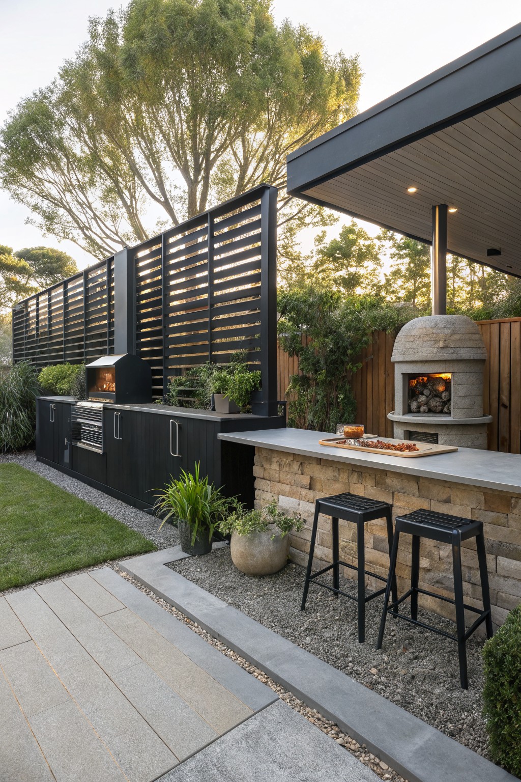 An outdoor kitchen with a tall black horizontal slat privacy screen behind dark cabinets, a stone pizza oven, and two black stools at a concrete bar.