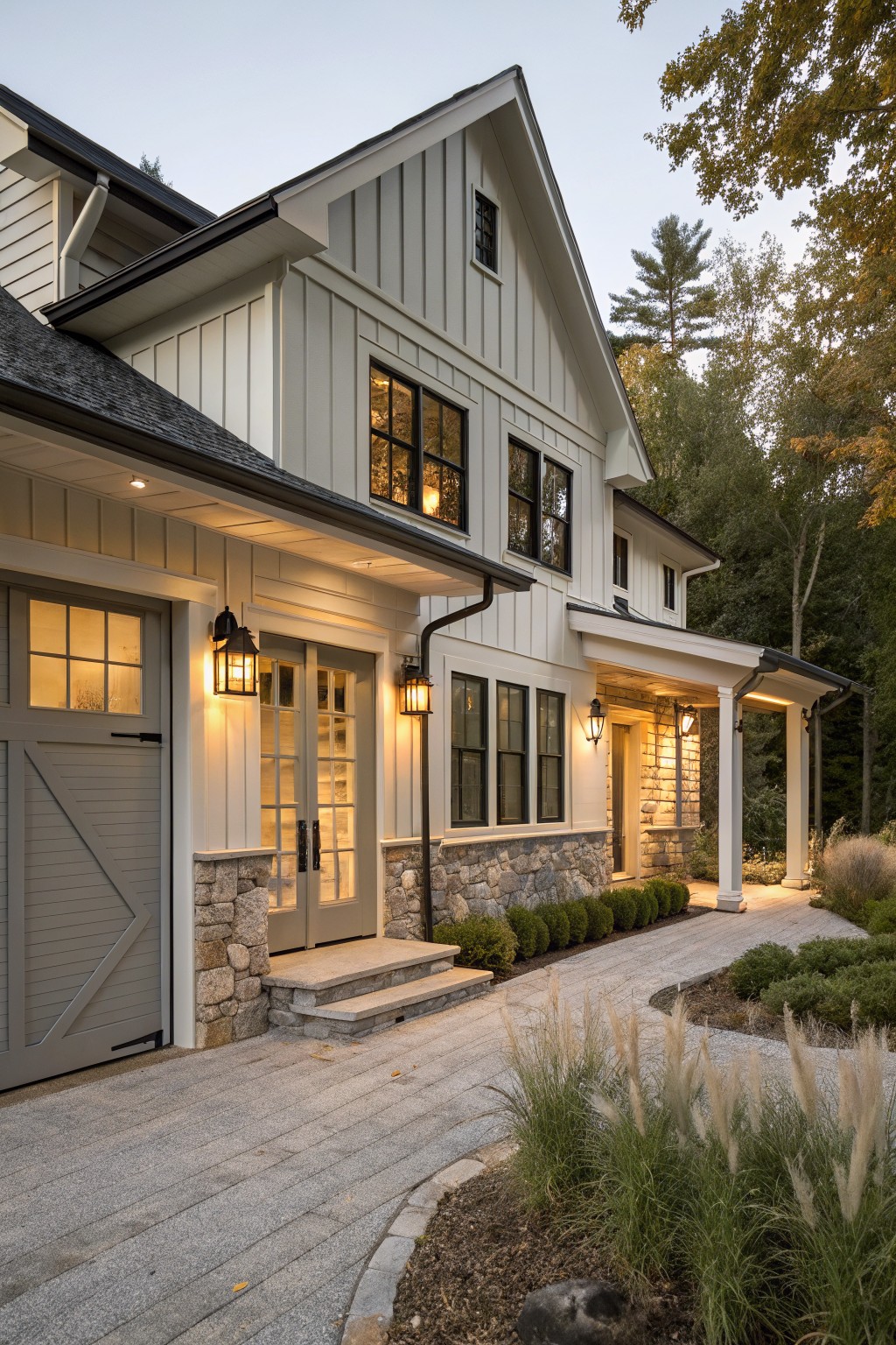 Modern white farmhouse with stone accents, lit windows, and landscaped driveway at dusk.
