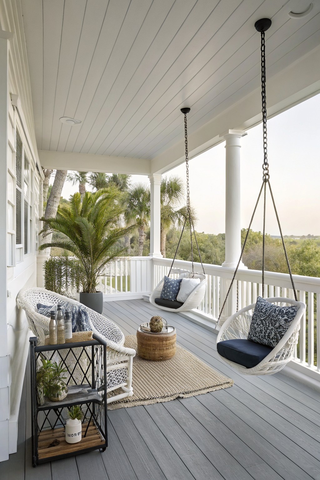 A white screened porch with two hanging white chairs, a wicker armchair, a jute rug, and gray decking overlooking trees.