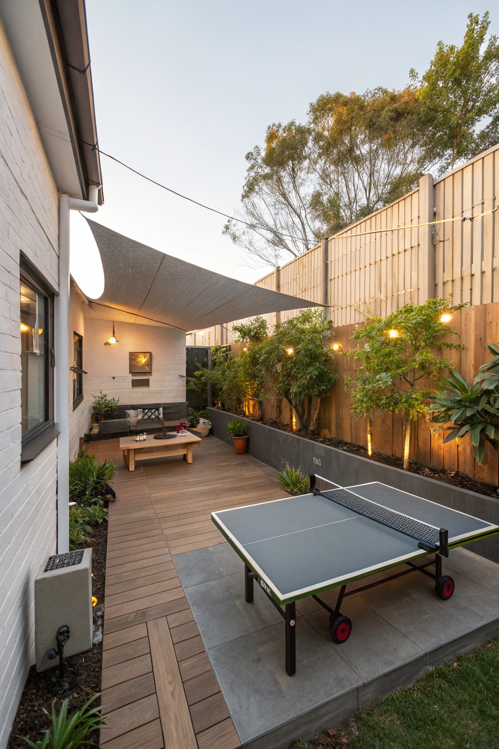 Backyard deck with a large gray shade sail overhead, built-in seating, wooden coffee table, and ping pong table on an adjacent concrete pad.