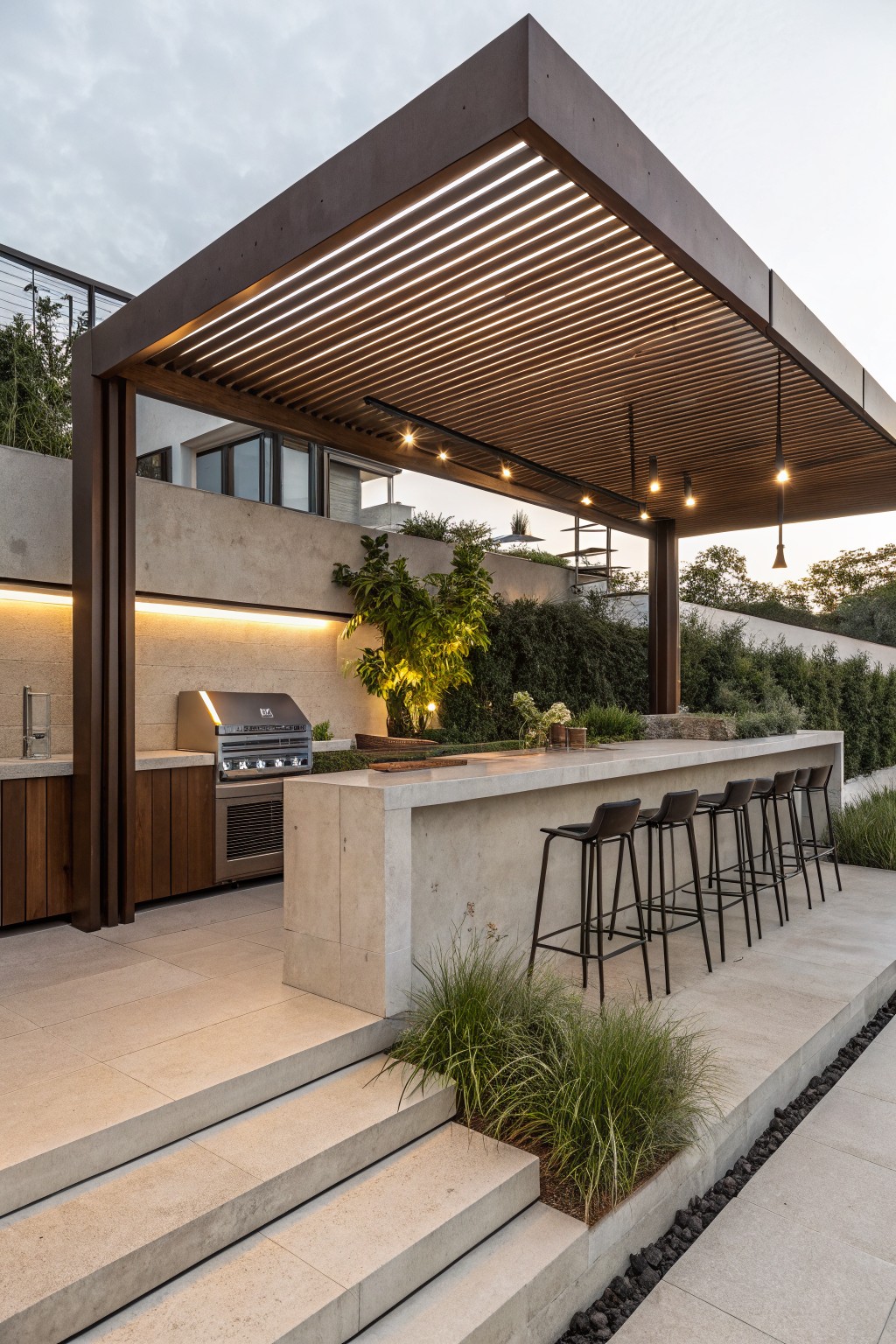 An outdoor patio with a long concrete bar counter lined with dark bar stools beneath a slatted wooden pergola that includes integrated lighting and an adjacent built-in grill area.