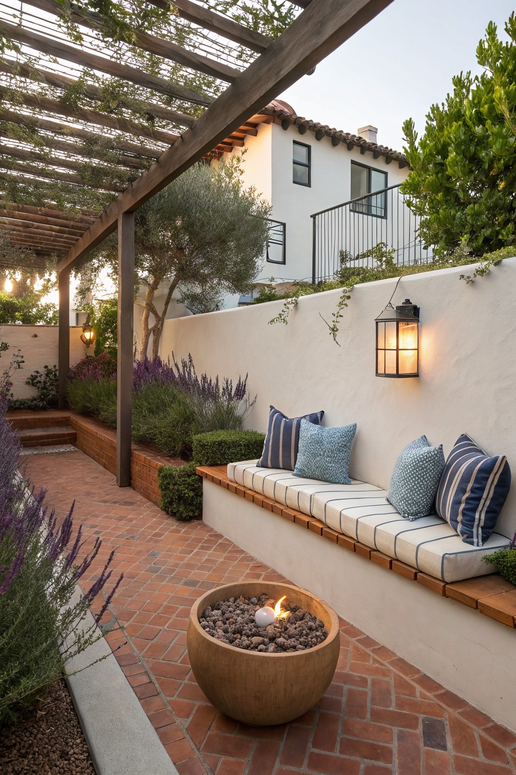 A compact brick patio with a wooden pergola overhead, built-in bench seating with blue and white cushions along a white stucco wall, and a round concrete fire pit in the center.