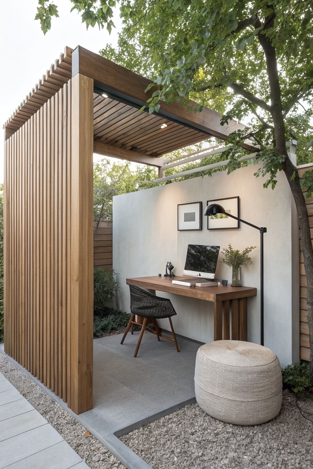An outdoor workspace under a wooden pergola with vertical slat screening on one side, featuring a desk, chair, and floor lamp against a concrete wall.