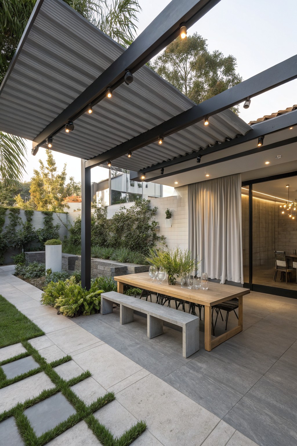 Outdoor patio under a dark metal pergola with a long wooden table, concrete bench, white side curtains, and surrounding garden plants.