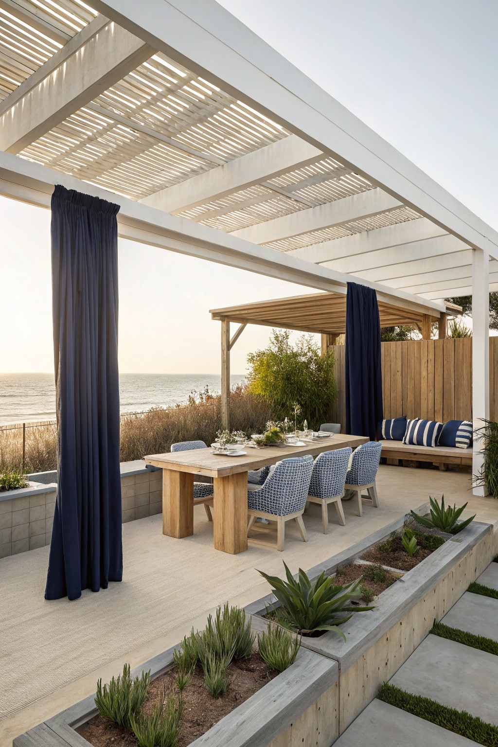A white pergola with blue side curtains over a wooden dining table and chairs on a coastal patio.