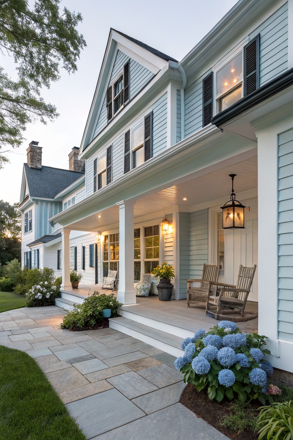 Light blue house with white porch, rocking chairs, and blue hydrangeas at dusk.