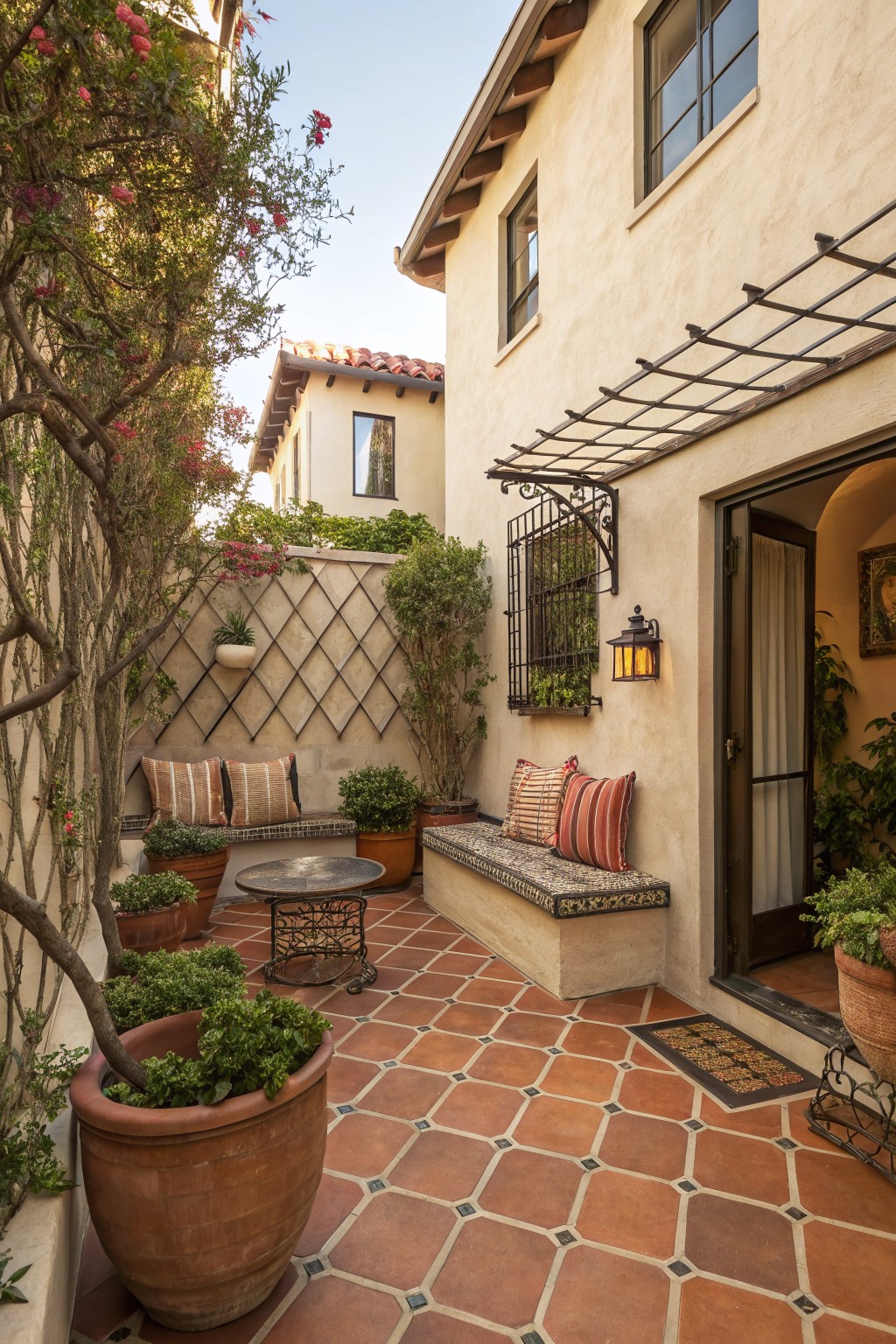 A compact courtyard with terracotta tile flooring, built-in stucco benches with cushions, potted plants, and an overhead metal trellis beside a stucco building.