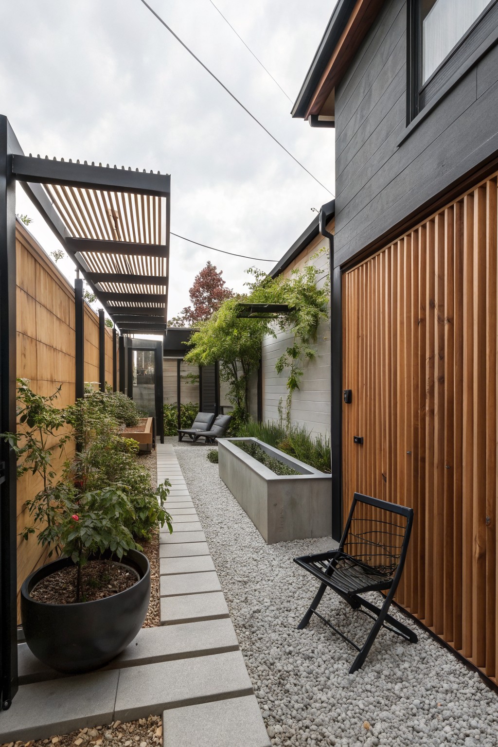 Narrow townhouse side yard with gravel path, concrete trough planter, black pergola, wooden fencing, and layered green plants.
