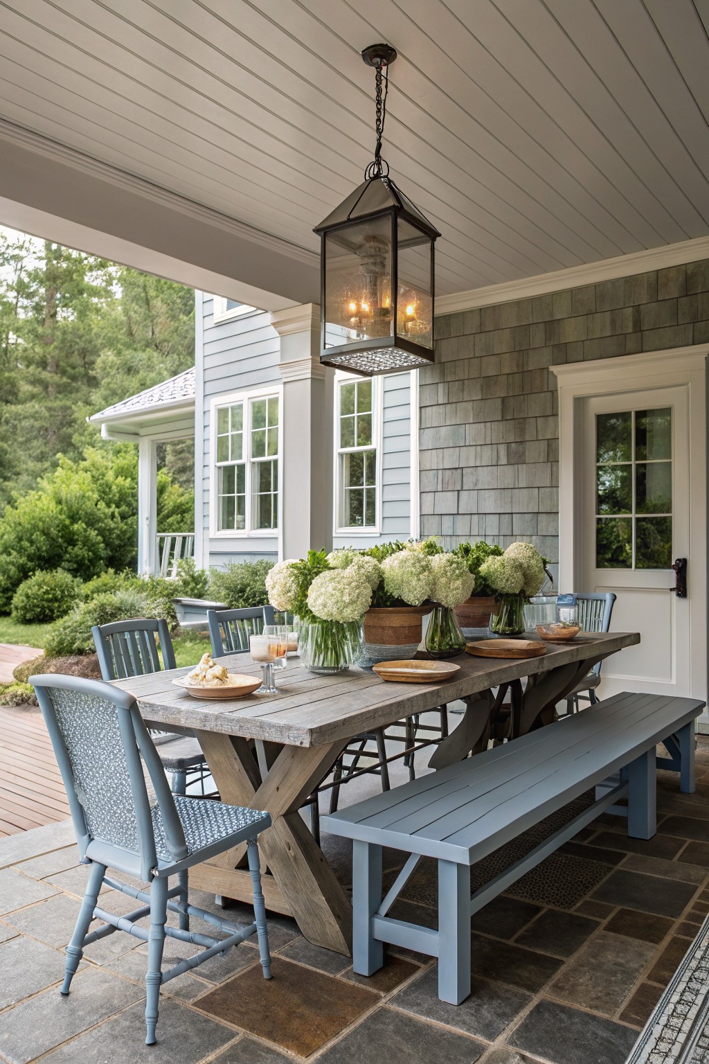 A long wooden dining table with blue chairs and a bench on one side sits on a stone patio under a covered porch with a large hanging lantern and white flowers in glass vases.