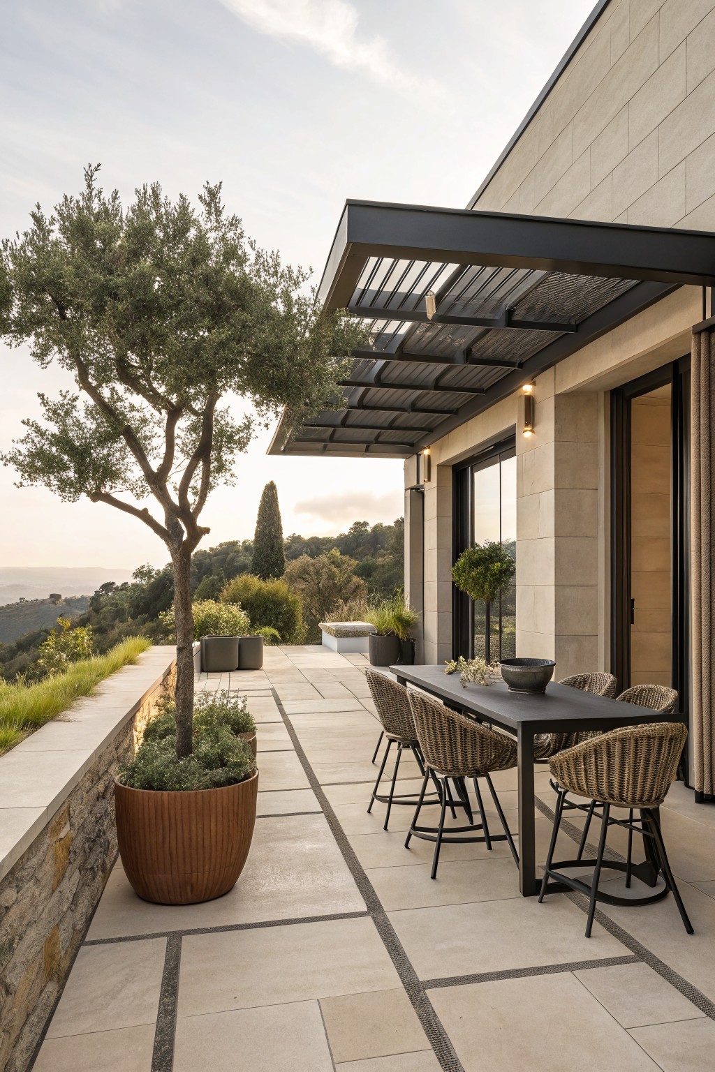 A modern outdoor patio features a long dining table with woven chairs beneath a black metal slatted roof structure, large terracotta pots with trees and shrubs along a stone wall, and a view of hills in the background.