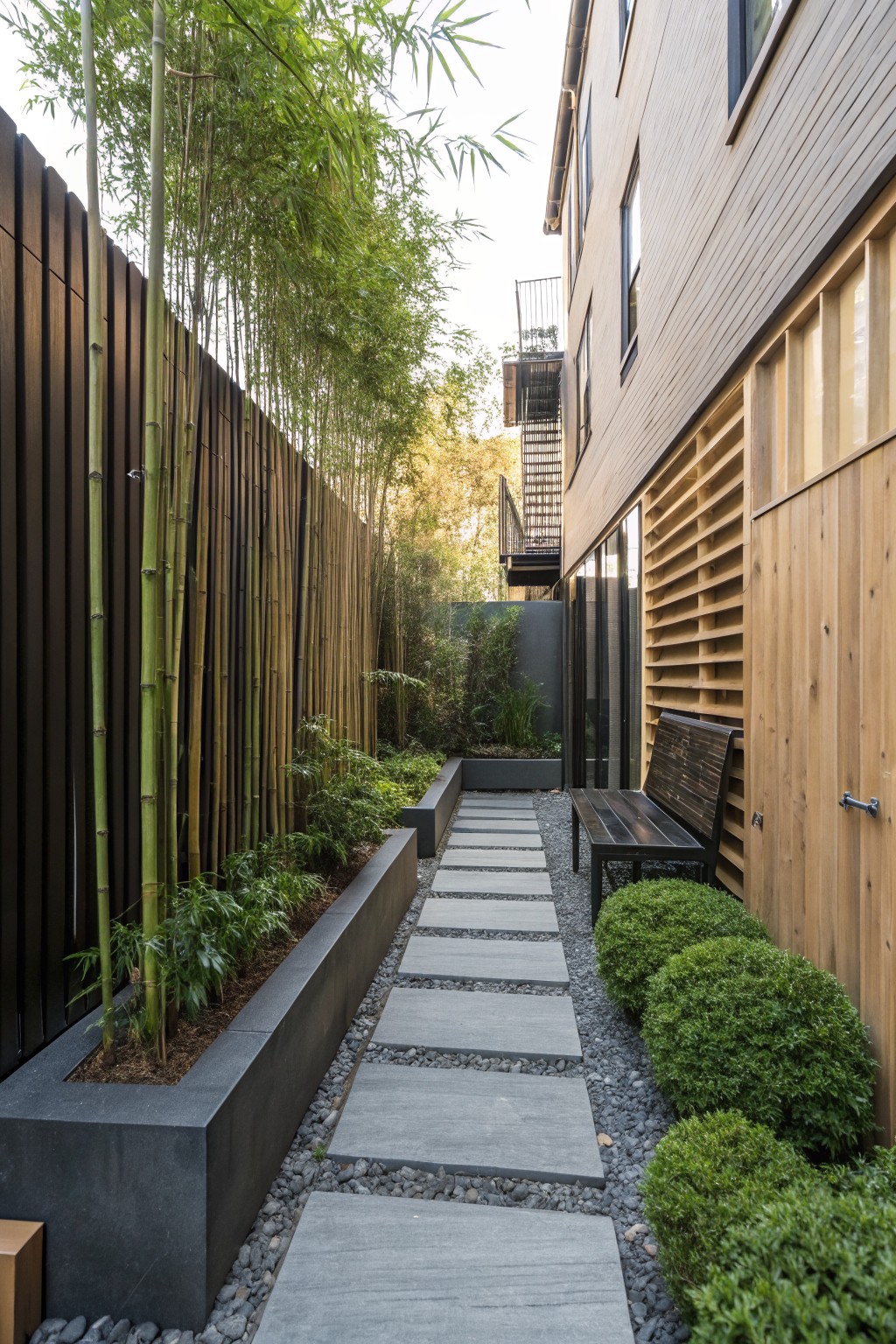 A narrow gravel path with gray stepping stones runs between tall bamboo plants on the left and a wooden building on the right, with low green shrubs along the edges.