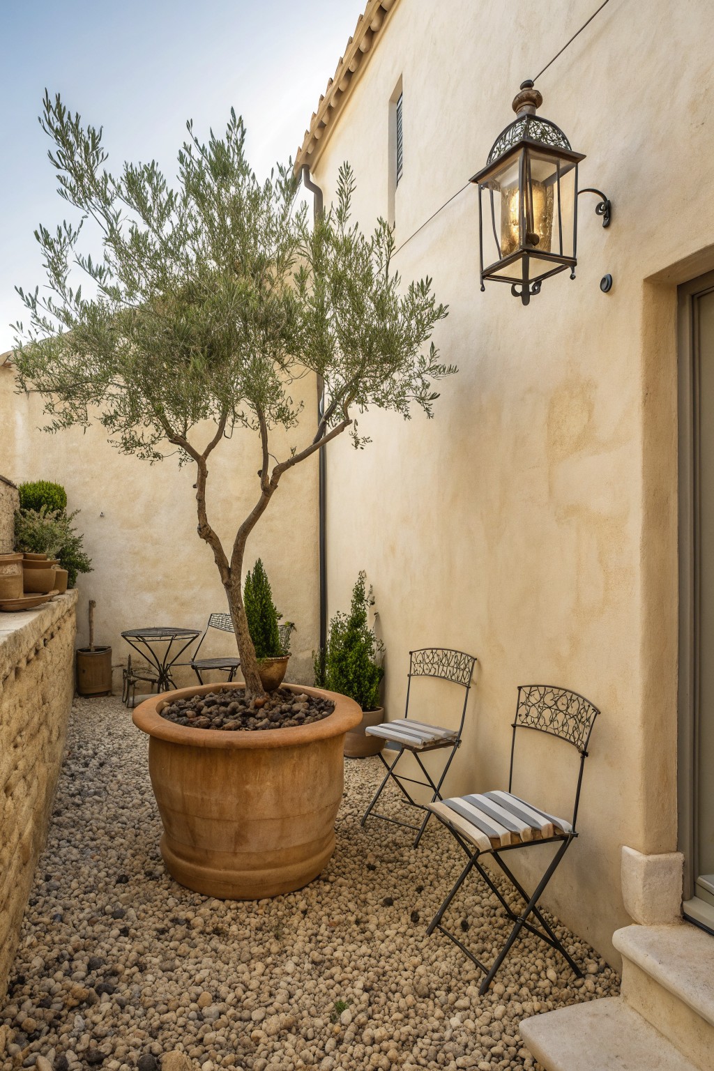 Courtyard with potted olive tree, striped chairs, and lit wall lantern