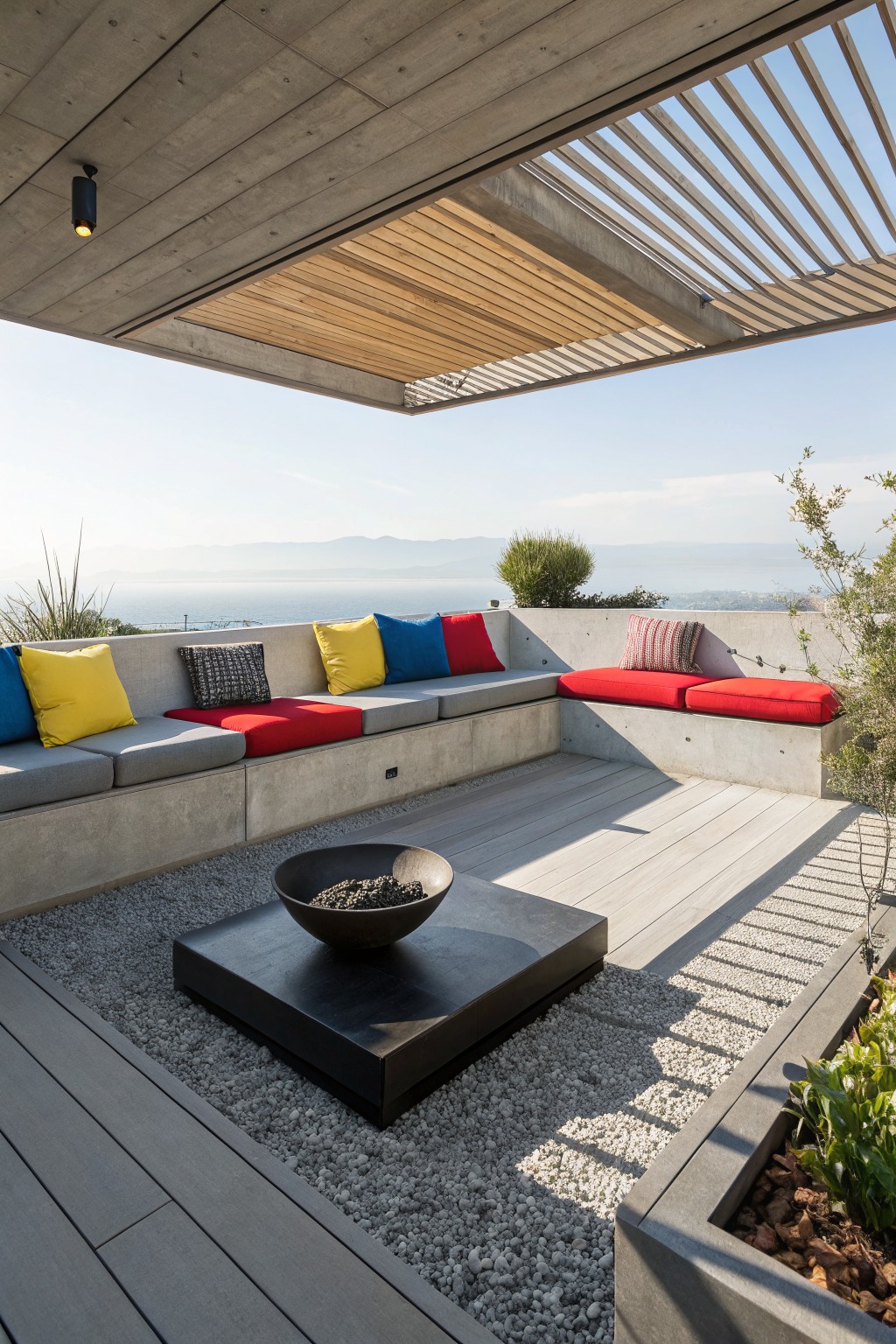 A modern outdoor terrace with L-shaped concrete built-in seating, colorful cushions in red, yellow, and blue, a black fire bowl on gravel, and wood decking with distant water views.