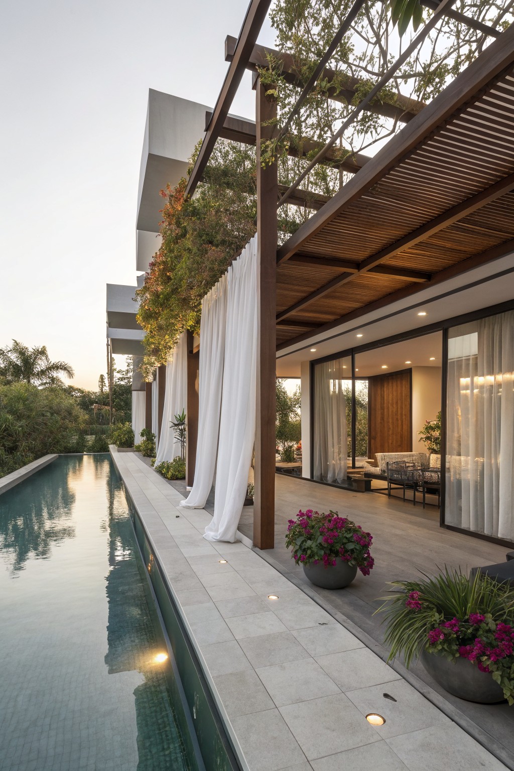 A modern outdoor patio with a wooden pergola, white sheer curtains, potted plants, and a long swimming pool beside a tiled deck.