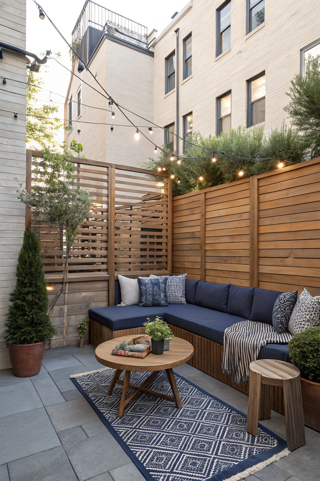 Backyard patio with navy built-in seating along a horizontal wood fence, a round coffee table on a patterned rug, and string lights overhead.