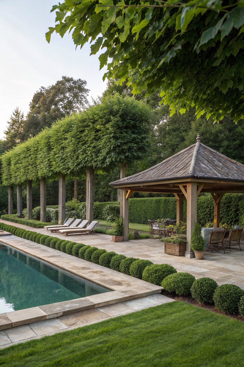 A backyard pool with a stone pavilion, lounge chairs, and tall green hedges lining the pool edge.