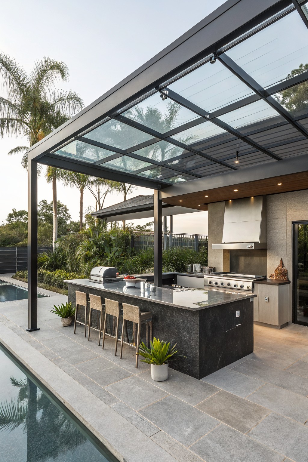 Outdoor kitchen with a long dark island, four bar stools, stainless appliances, and a glass roof structure next to a swimming pool.