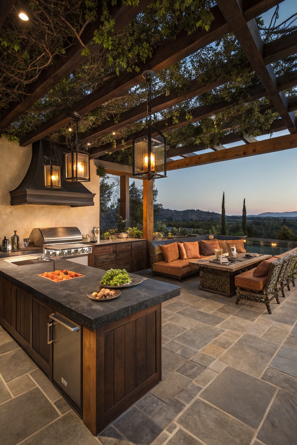 An outdoor kitchen patio with a large dark stone island, stainless steel grill, wooden cabinetry, and lounge seating under a vine-covered pergola overlooking a landscape at dusk.