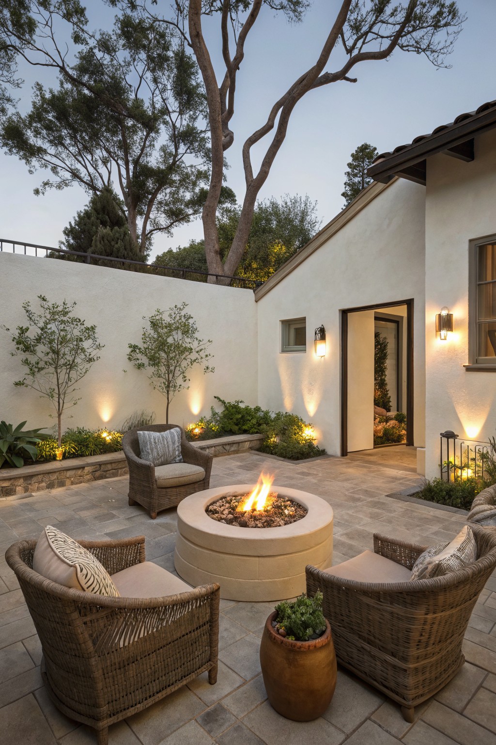 A round concrete fire pit sits at the center of a stone patio surrounded by four wicker armchairs, with a white stucco house wall, open doorway, and trees in the background.