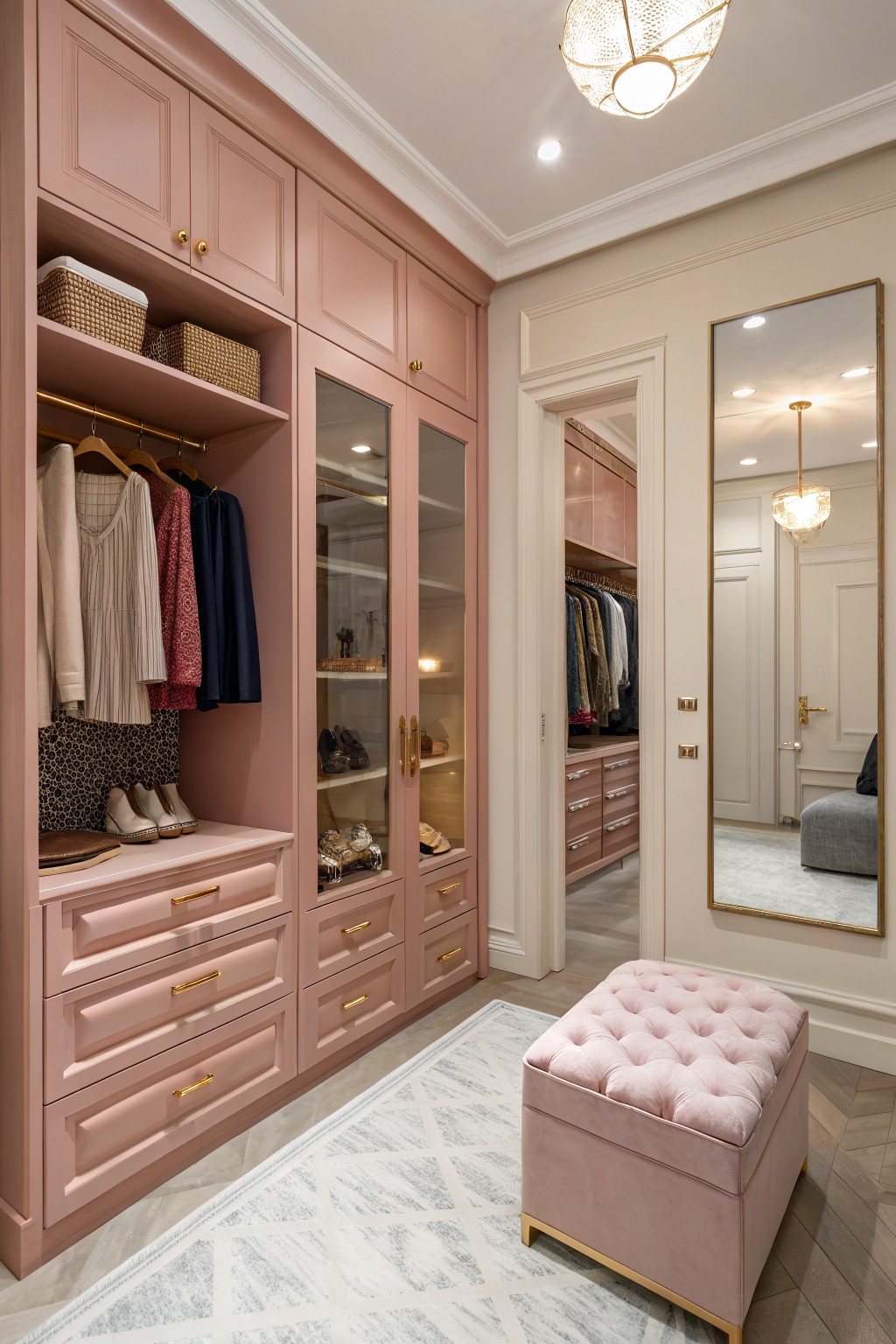 Pink walk-in closet with glass cabinets, hanging clothes, shoes, and tufted ottoman.