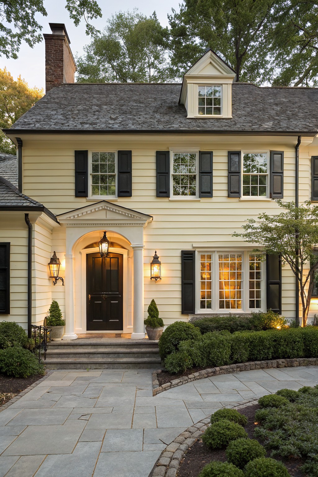 A pale yellow house exterior with dark shutters and stone paving.