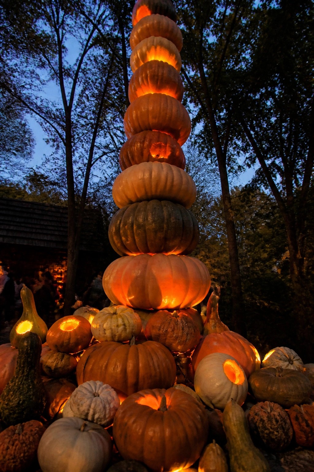 Tall outdoor stack of carved and lit pumpkins forming a glowing tower surrounded by more pumpkins at the base.
