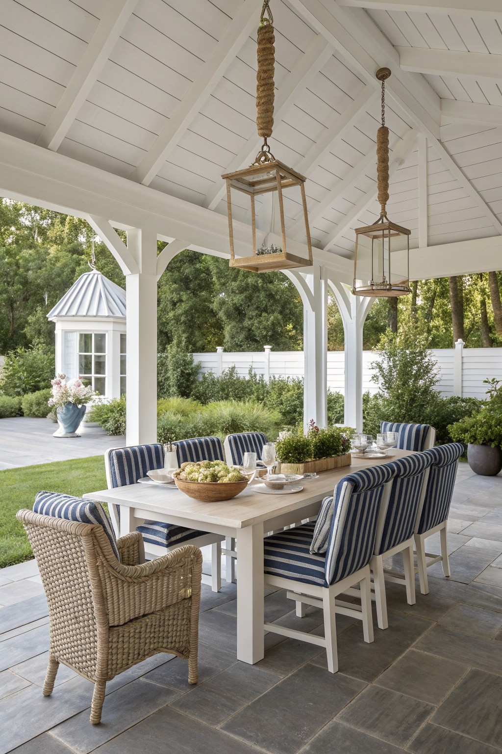 A long white dining table with blue striped chairs and one woven chair sits under a white gazebo with hanging lanterns.