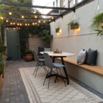 A narrow outdoor patio features a long built-in wooden bench with cushions along a concrete wall, a small square table with two modern chairs, potted plants, and string lights overhead.