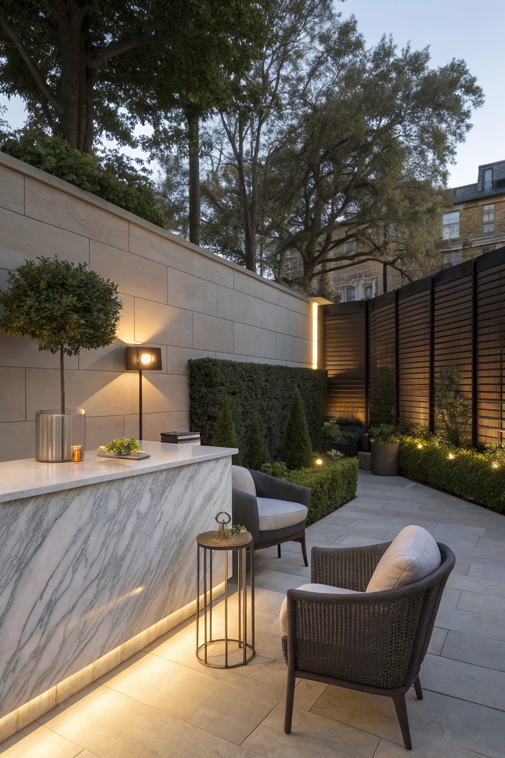 A narrow outdoor patio featuring a long marble counter along a stone wall, two armchairs, a round side table, and soft wall lighting.
