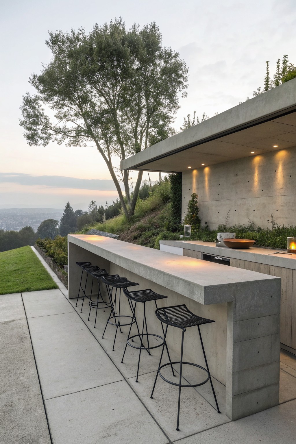 An outdoor concrete bar counter with black metal stools lines a modern patio overlooking a hillside.