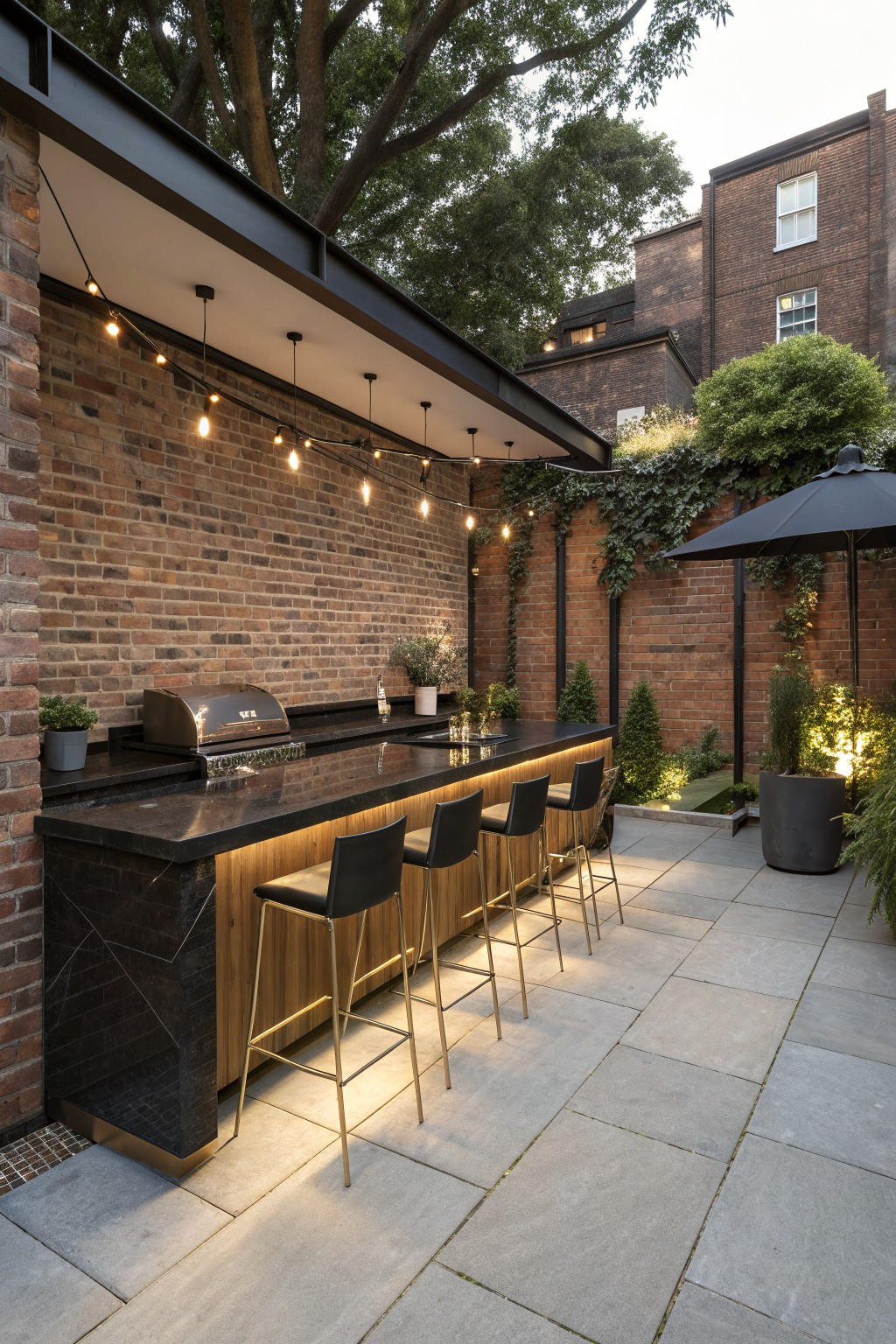 Outdoor bar with a long dark countertop and row of black counter-height stools on a paved patio next to a brick wall.