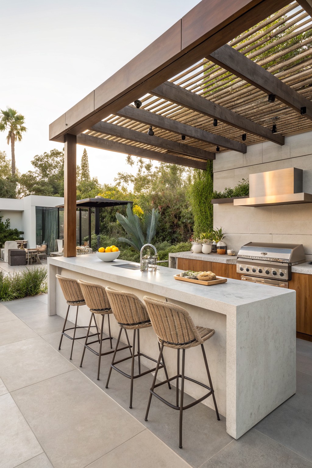 Outdoor kitchen bar with four woven counter-height stools along a concrete counter under a wooden pergola, grill visible on the right.