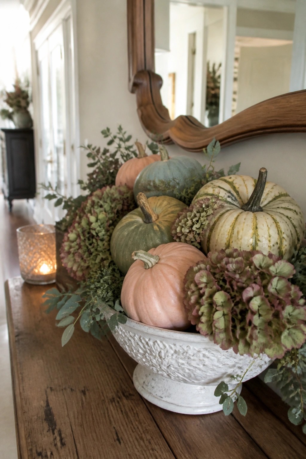 A white bowl on a wooden console holds several whole pumpkins in peach, green, and striped white tones, surrounded by dried hydrangeas and greenery beneath a large mirror.