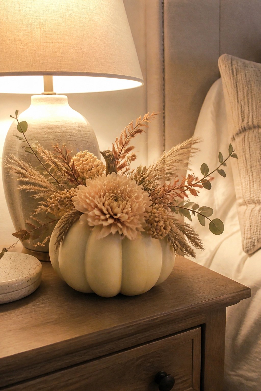A white pumpkin filled with dried beige flowers and grasses sits on a wooden nightstand beside a lit lamp.