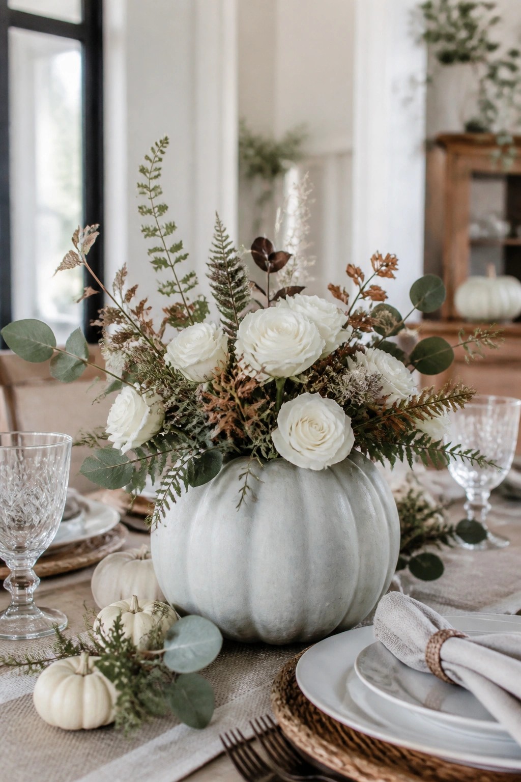 Light gray painted pumpkin used as a vase holding white roses and mixed greenery as the main centerpiece on a set dining table.