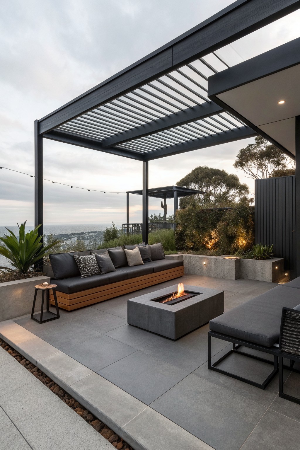 A modern outdoor patio with a long built-in bench featuring dark cushions and a wood base under a dark slatted pergola, a rectangular concrete fire pit in the center, and a view toward the ocean at dusk.