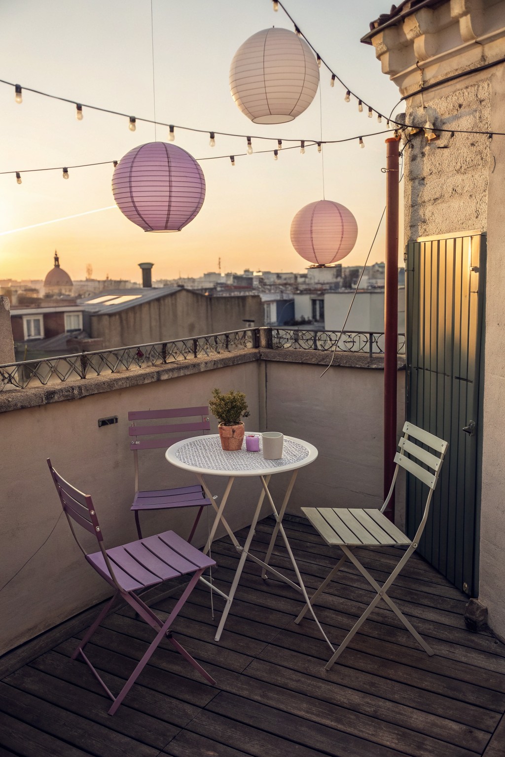 A small urban rooftop terrace with a round white table, three folding chairs in purple and white, a potted plant, and several hanging paper lanterns connected by string lights overlooking city buildings at sunset.
