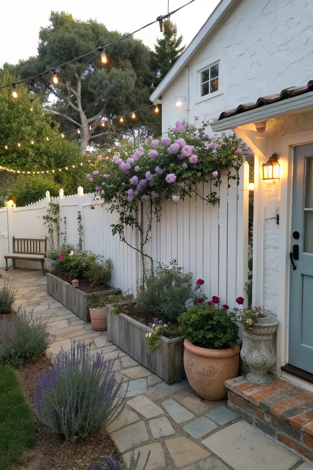 A white wooden fence with built-in raised planters holding pink roses and other flowers, next to a stone path and house entrance.