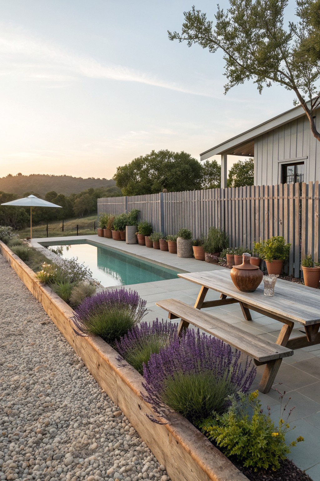 A rectangular pool next to a tall wooden fence lined with rows of potted plants and a wooden picnic table on the stone patio.