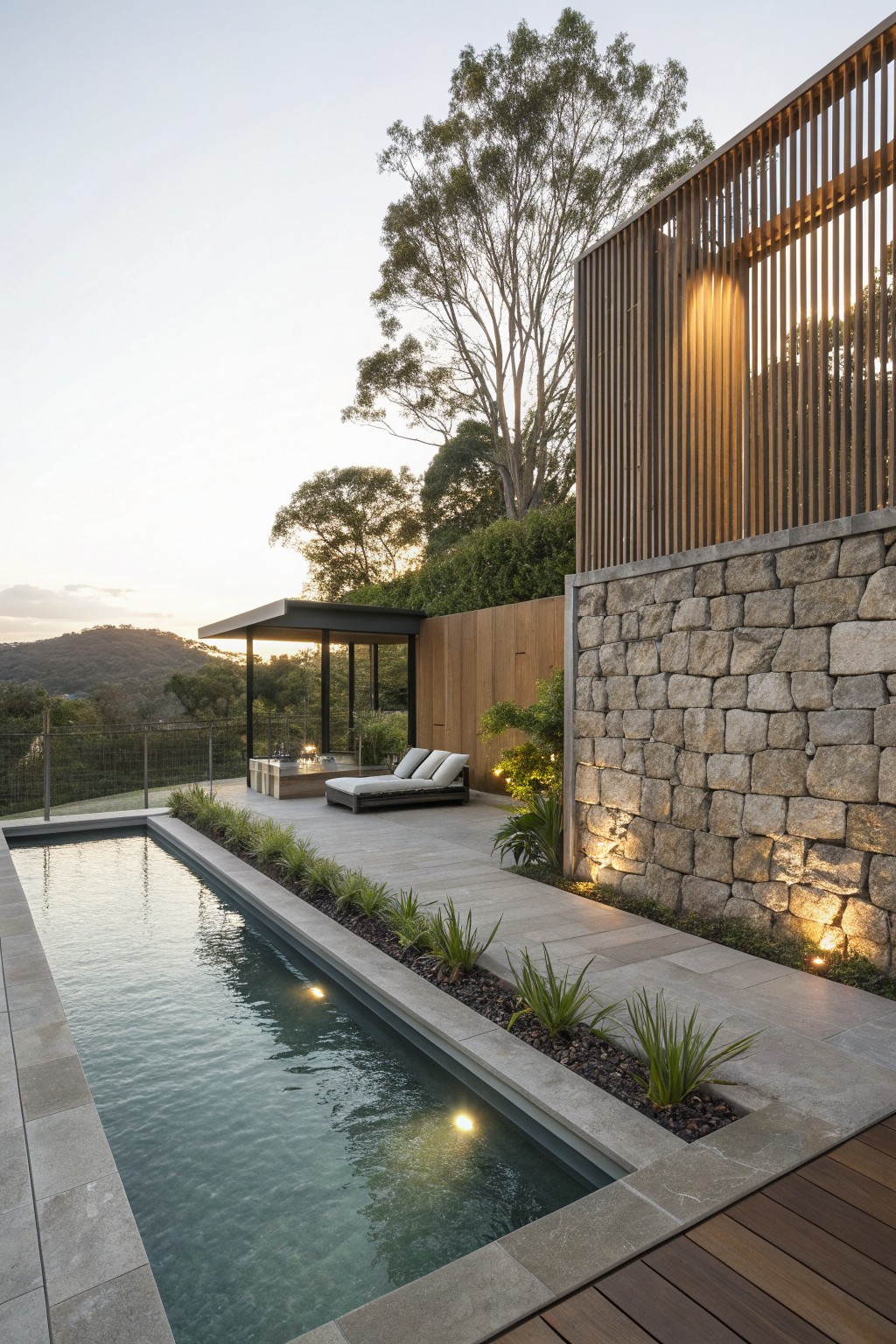 Modern infinity pool with lounge chairs beside illuminated stone wall at sunset